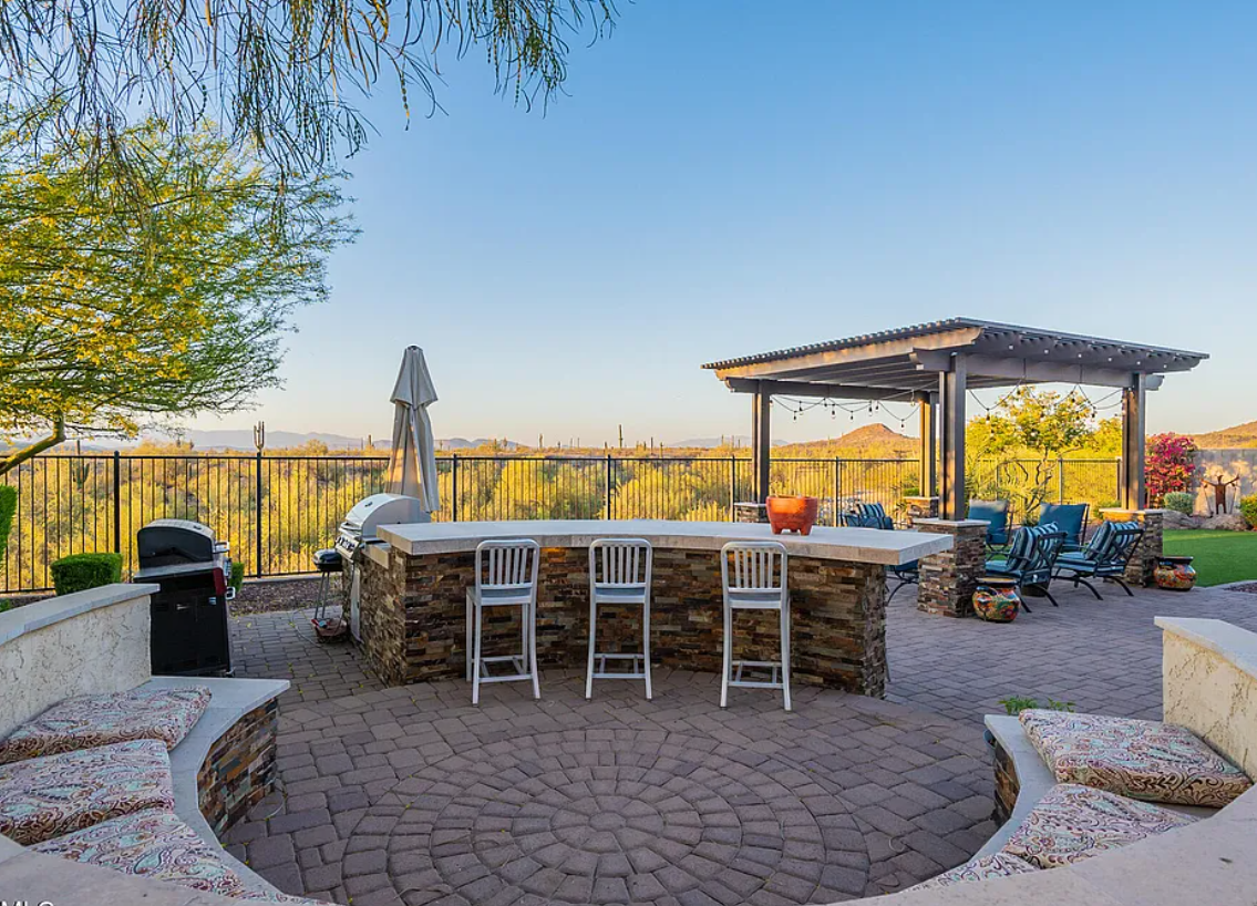 Resort-style backyard at 9934 W White Feather Ln — outdoor kitchen with bar seating, pergola lounge, and desert mountain views